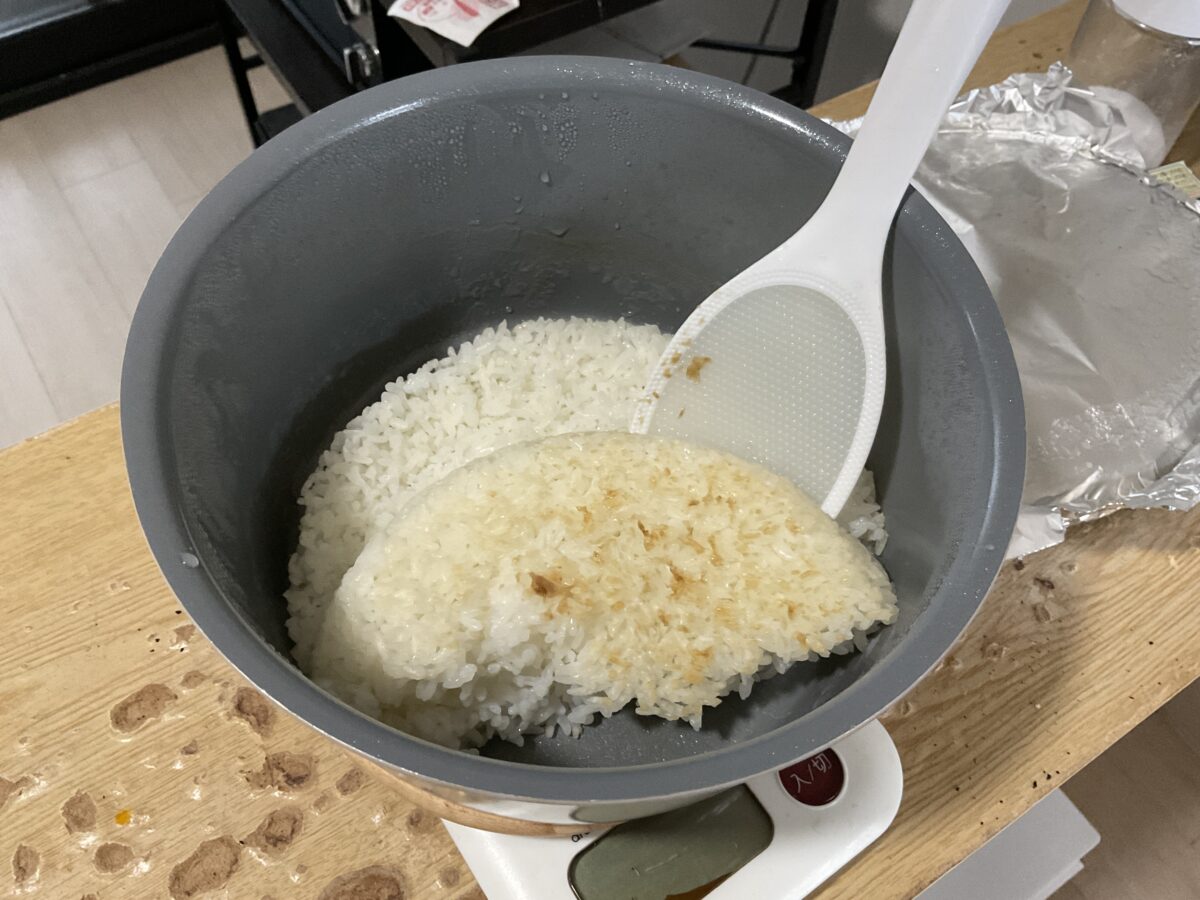 一人暮らしのショウが鍋で炊いたご飯をしゃもじですくいおこげを確認している様子